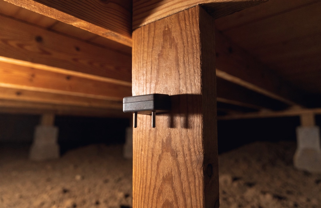 A pin-type moisture meter pressed against a wooden support post in a crawl space, with floor joists and masonry piers visible behind